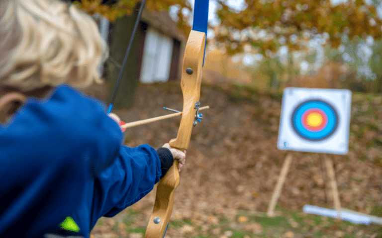 Children and teenagers practicing archery in a forest clearing. Target stands set up at varying distances. Trees and foliage in the background