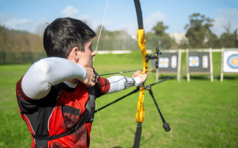 An archer draws back their bow, focusing on the target ahead. The serene atmosphere of the traditional archery range contrasts with the precision and competitiveness of the Olympic archery range