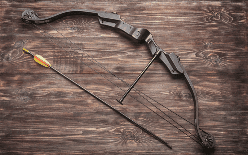 A bow and arrow lie on a wooden table next to a target with arrows stuck in it. A training plan for archers is visible in the background